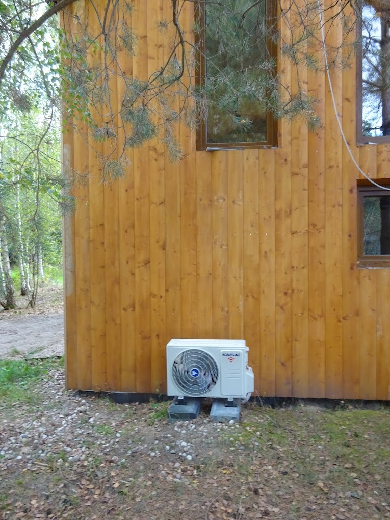 Biała jednostka zewnętrzna klimatyzacji Kaisai, zamontowana przy drewnianej elewacji budynku. Widoczne okna i otoczenie leśne. Element instalacji na zewnątrz budynku.