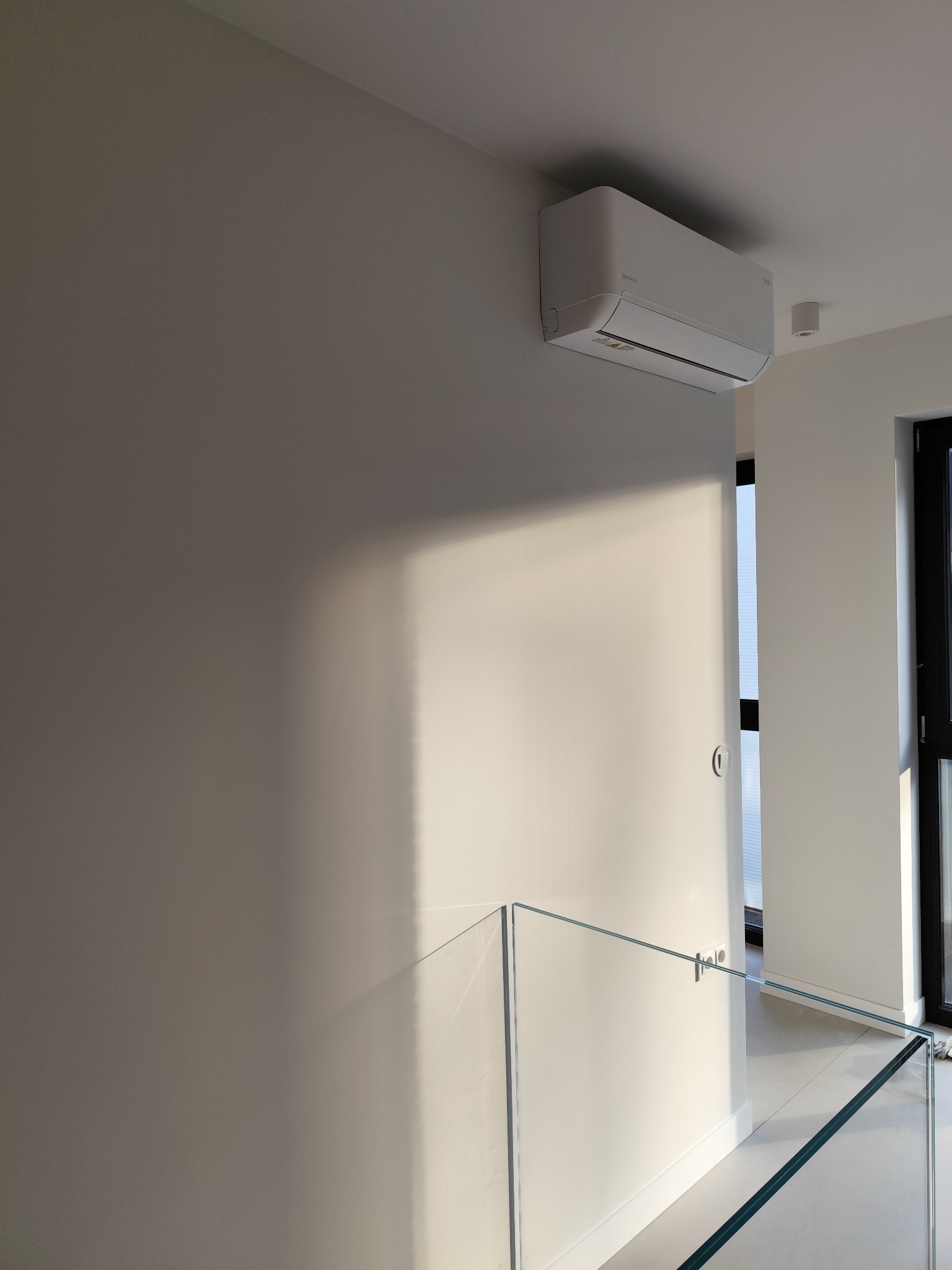 Nowoczesny, biały klimatyzator ścienny zamontowany w minimalistycznym wnętrzu z szklaną balustradą i oświetleniem punktowym. Widoczne światło słoneczne na ścianie.