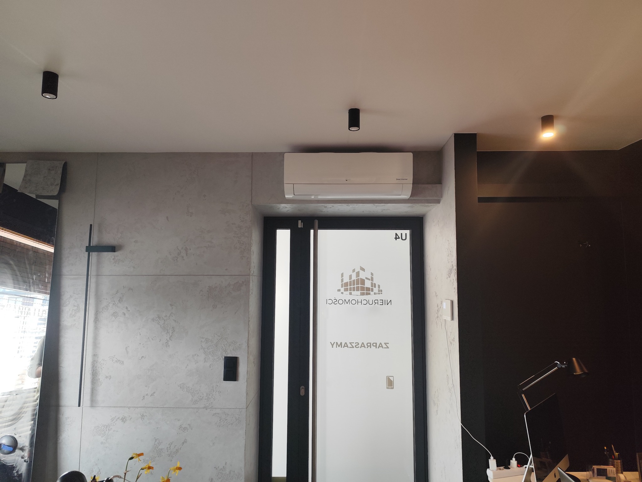 Nowoczesne wnętrze biurowe z klimatyzatorem LG na ścianie, widoczne oświetlenie punktowe i dekoracyjna ściana z efektem betonu.