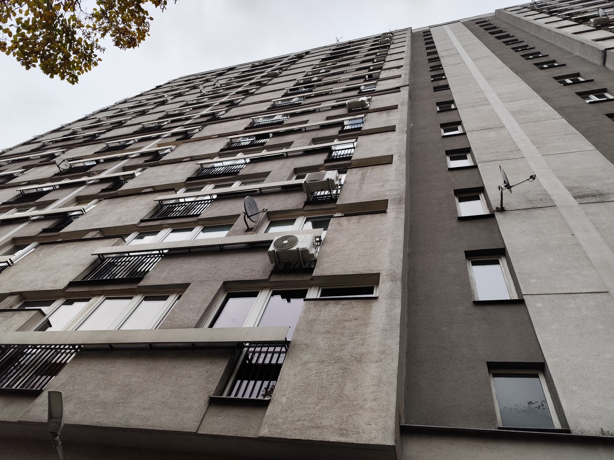 Szary blok z klimatyzatorami na ścianach, widok z dołu, akcent na urządzenia i architekturę budynku. Perspektywa podkreśla wysokość i powtarzalność elementów.