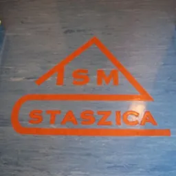 Niebieska podłoga z wykładziny z pomarańczowym logo 'ISM Staszica' w korytarzu.