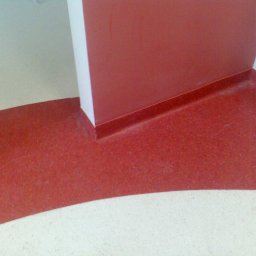 DĄBROWA GÓRNICZA - Fragment podłogi z wykładziną w odcieniach czerwieni, płynnie przechodząca w białą posadzkę w rogu pomieszczenia, przy ścianie pomalowanej na czerwono.