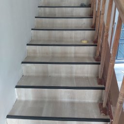 DĄBROWA GÓRNICZA - Schody wykończone panelami z drewnianą balustradą, widoczne czarne listwy zabezpieczające krawędzie stopni, taśma malarska na jednym ze stopni.