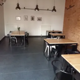 Wnętrze lokalu gastronomicznego z surowym wykończeniem: drewniane stoły z metalowymi nogami, czarne krzesła, szara posadzka, białe ściany z obrazkami i ceglana ściana po prawej stronie, oświetlenie...