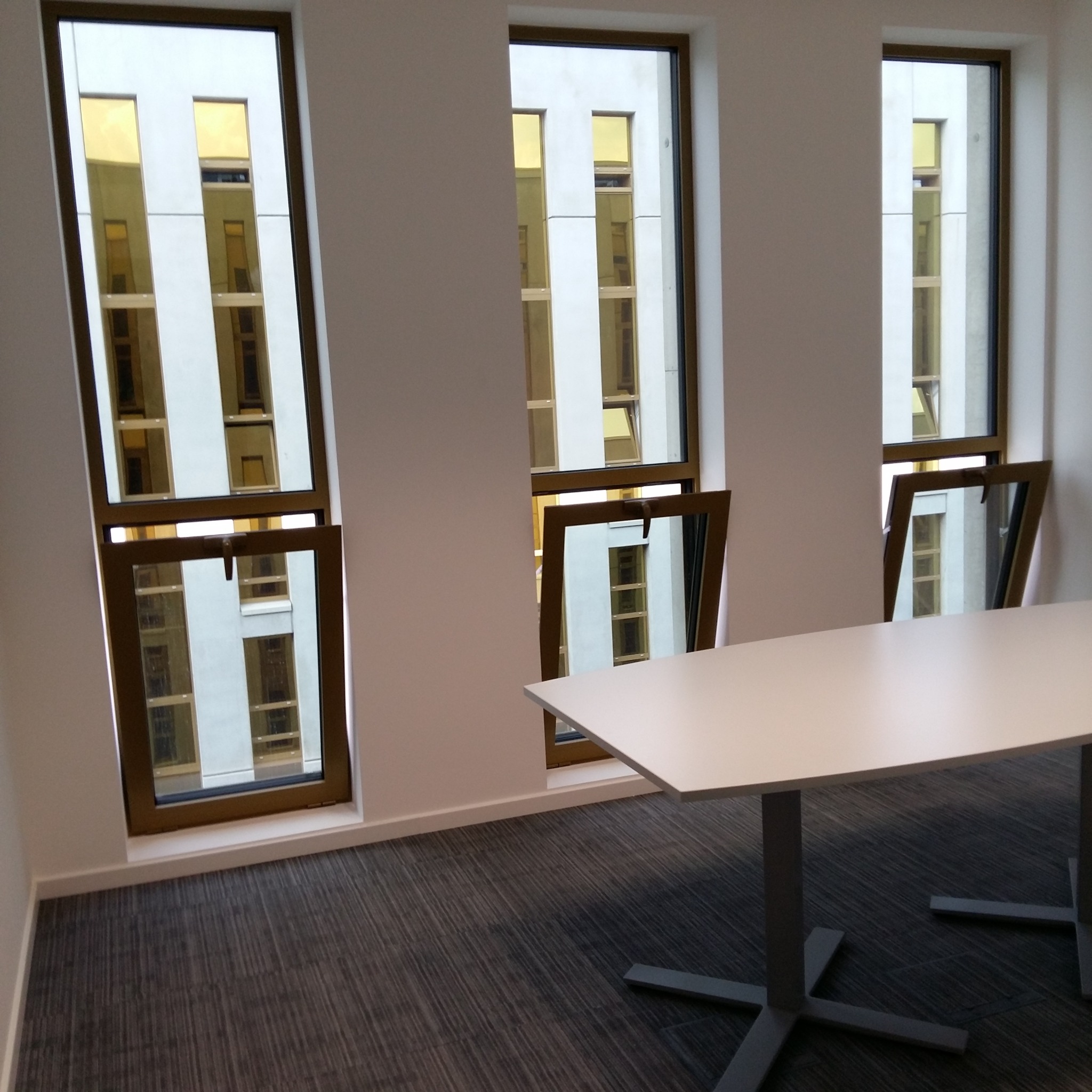 Jasne biuro z trzema oknami uchylnymi, szara wykładzina dywanowa i biały stół konferencyjny.