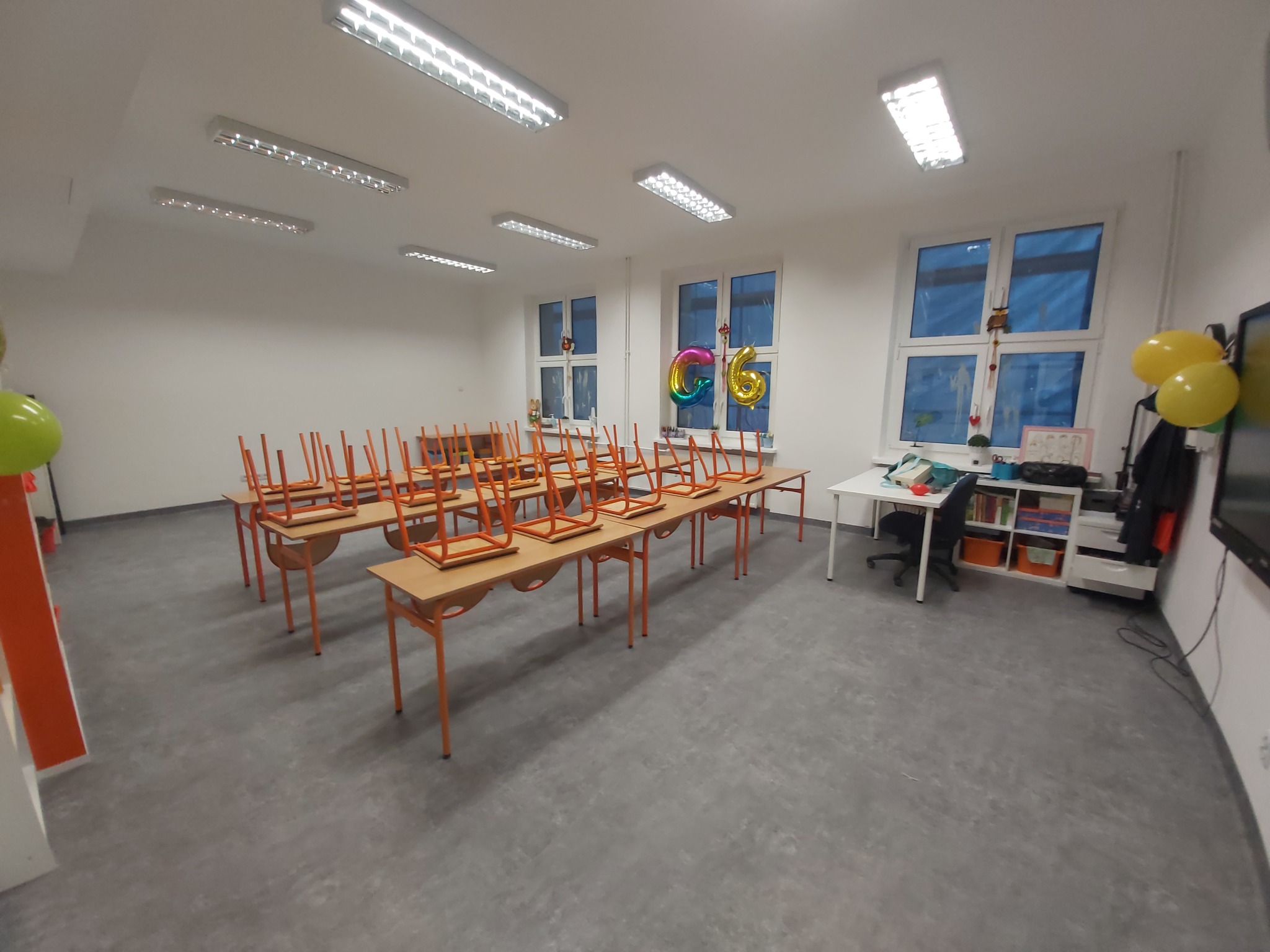 Sala lekcyjna z szarą podłogą, pomalowana na biało, z rzędami stolików z krzesłami na nich, przygotowana na uroczystość, o czym świadczą balony przy oknach i na ścianie.