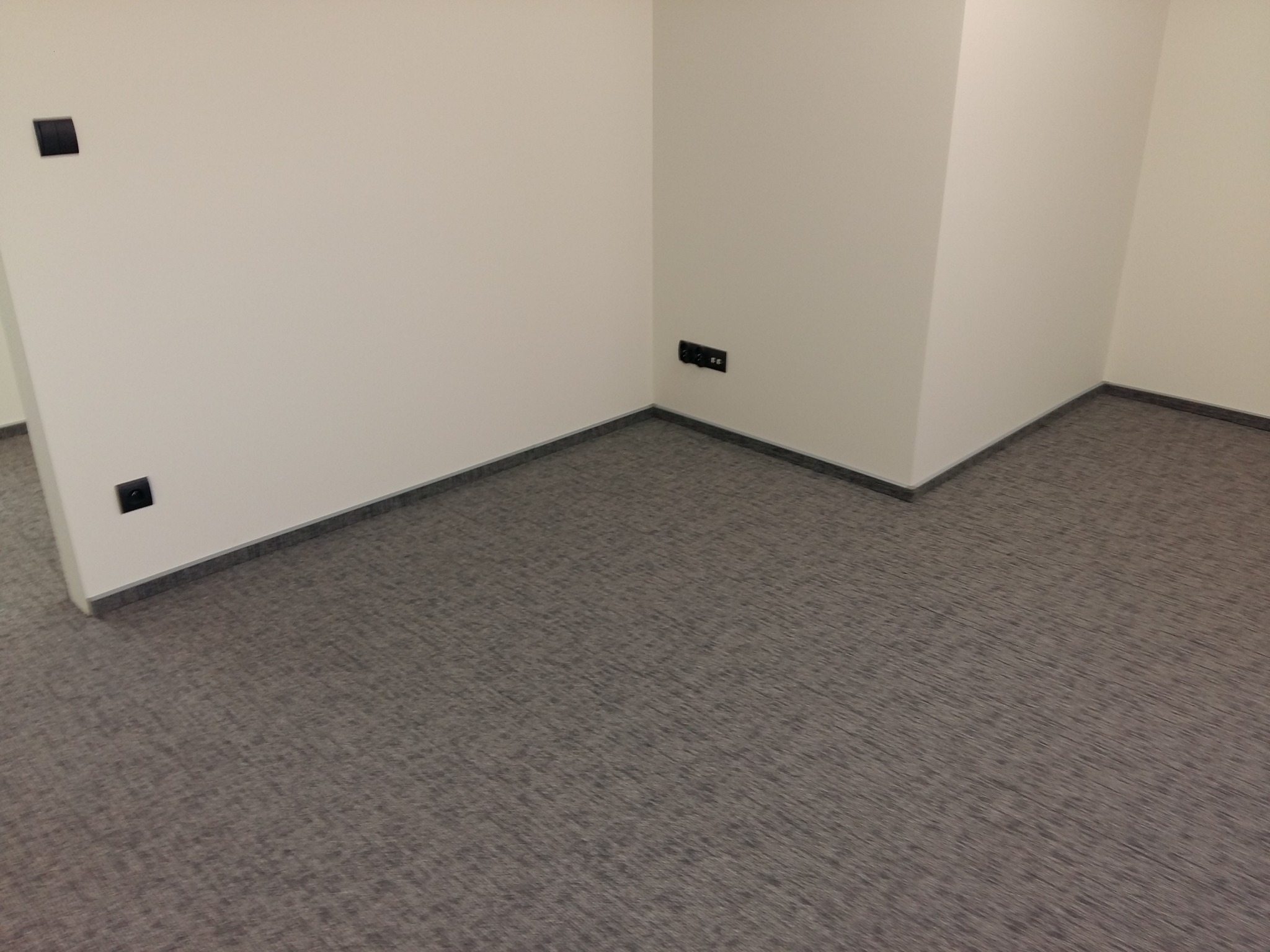 Szara wykładzina dywanowa w rogu jasnego pomieszczenia biurowego, widoczne czarne gniazdka elektryczne i włączniki światła.