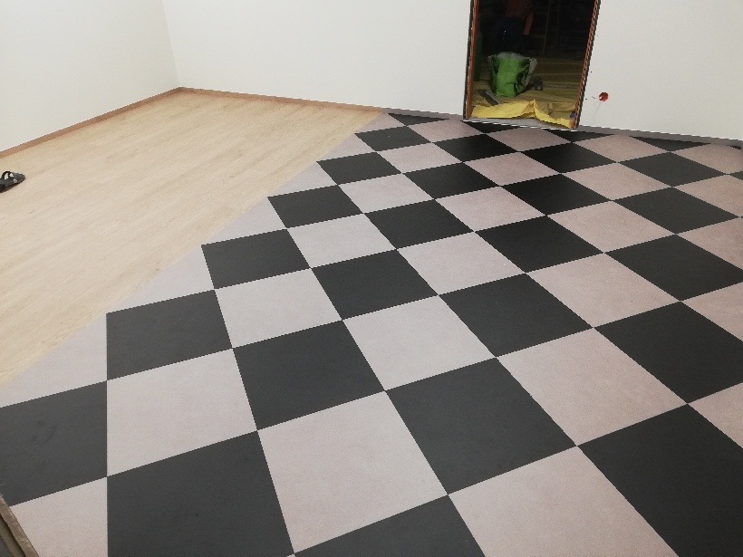 Fragment pokoju z dwiema różnymi podłogami: parkietem i wzorem szachownicy z płytek lub wykładziny, widoczne przejście między materiałami, niedokończona podłoga.