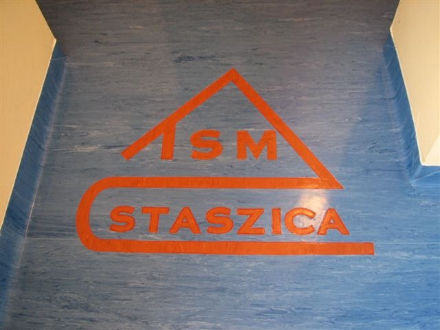 Niebieska podłoga z wykładziny z pomarańczowym logo 'ISM Staszica' w korytarzu.