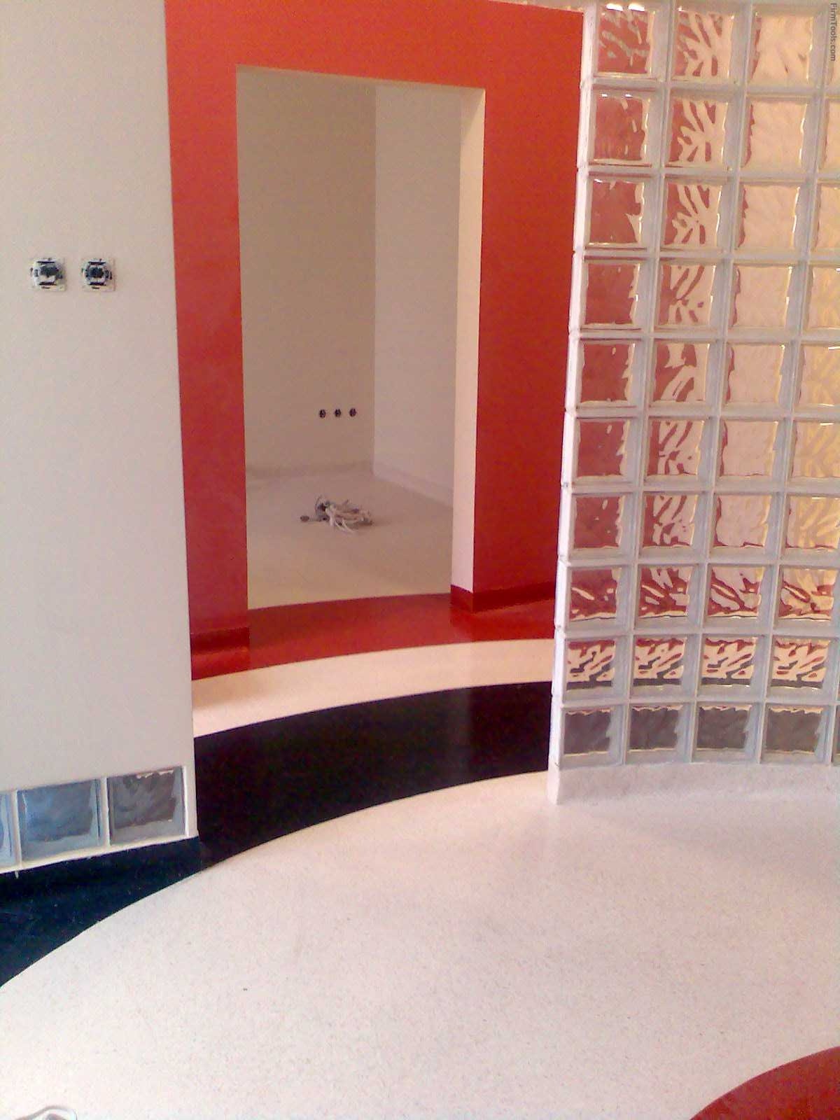 Wnętrze łazienki z dekoracyjną ścianką z luksferów, czerwonymi akcentami na ścianach i nietypową podłogą w pasy czarno-biało-czerwone, widoczne dwa gniazdka elektryczne na ścianie.