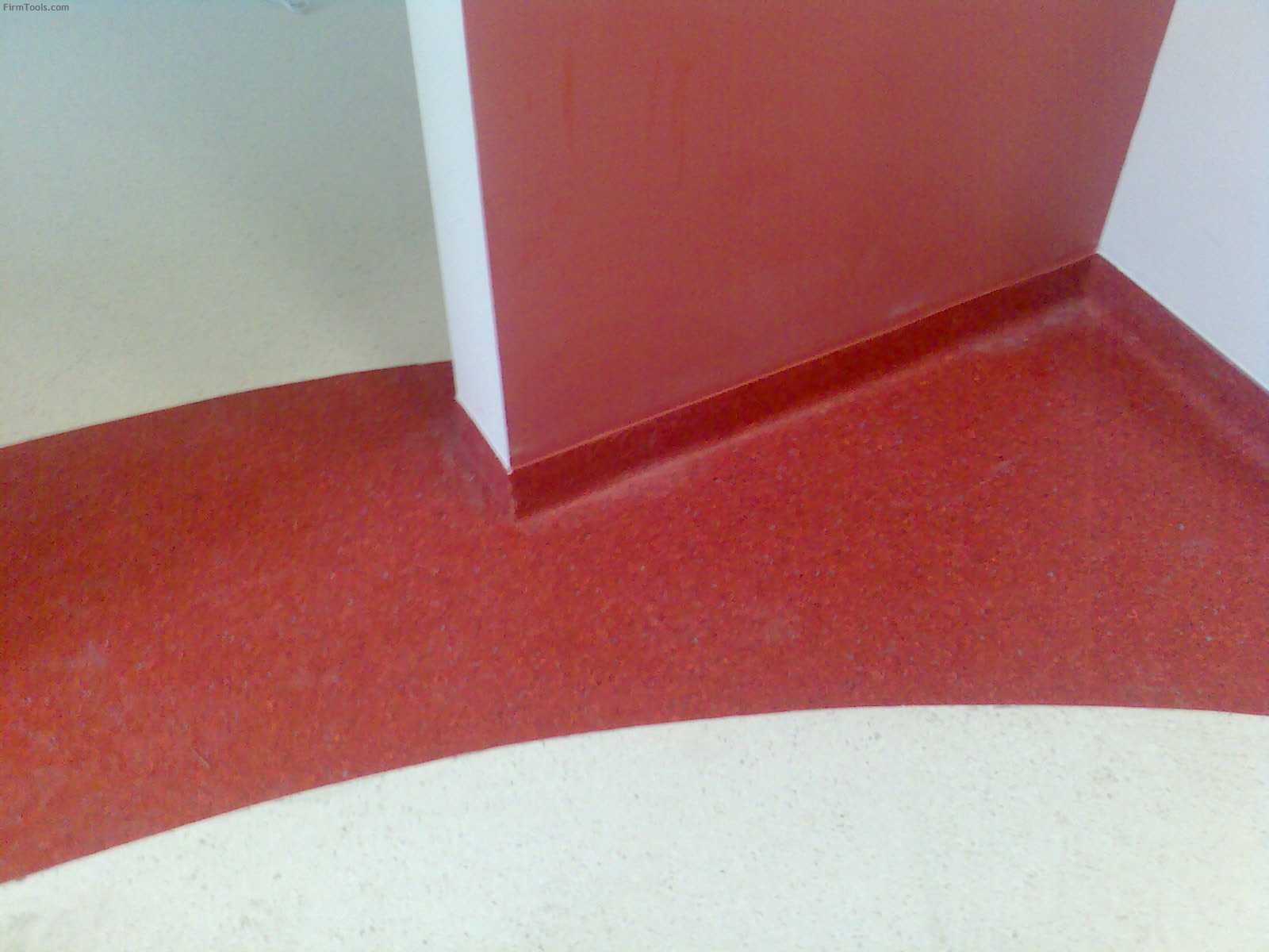 Fragment podłogi z wykładziną w odcieniach czerwieni, płynnie przechodząca w białą posadzkę w rogu pomieszczenia, przy ścianie pomalowanej na czerwono.