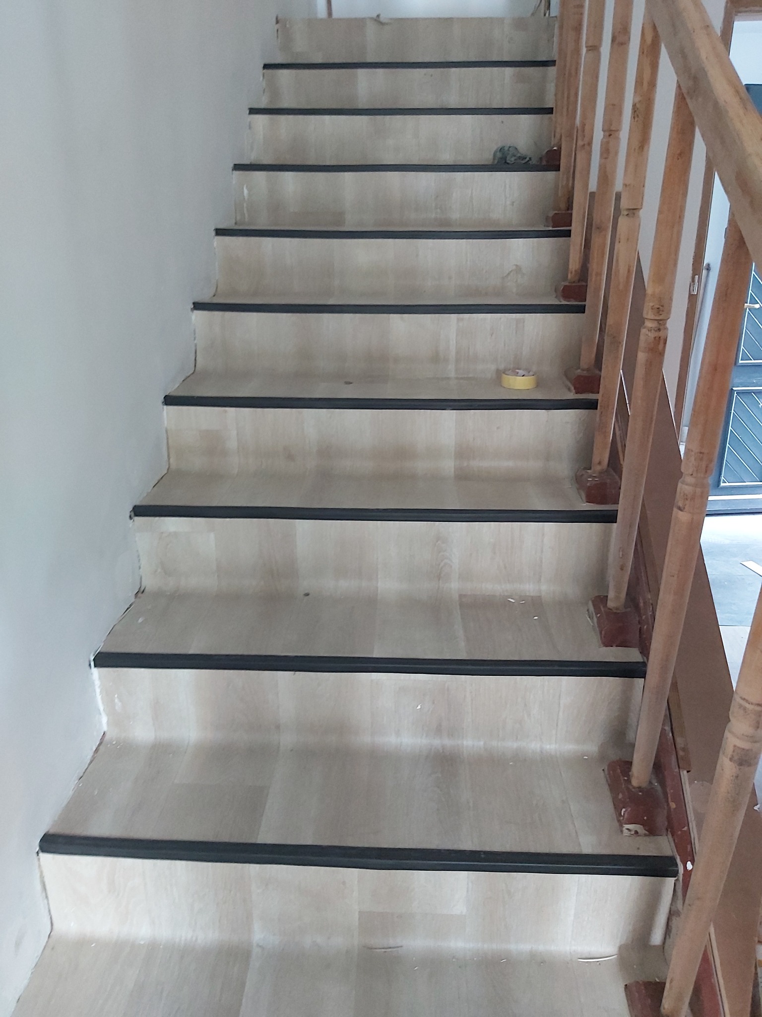 Schody wykończone panelami z drewnianą balustradą, widoczne czarne listwy zabezpieczające krawędzie stopni, taśma malarska na jednym ze stopni.