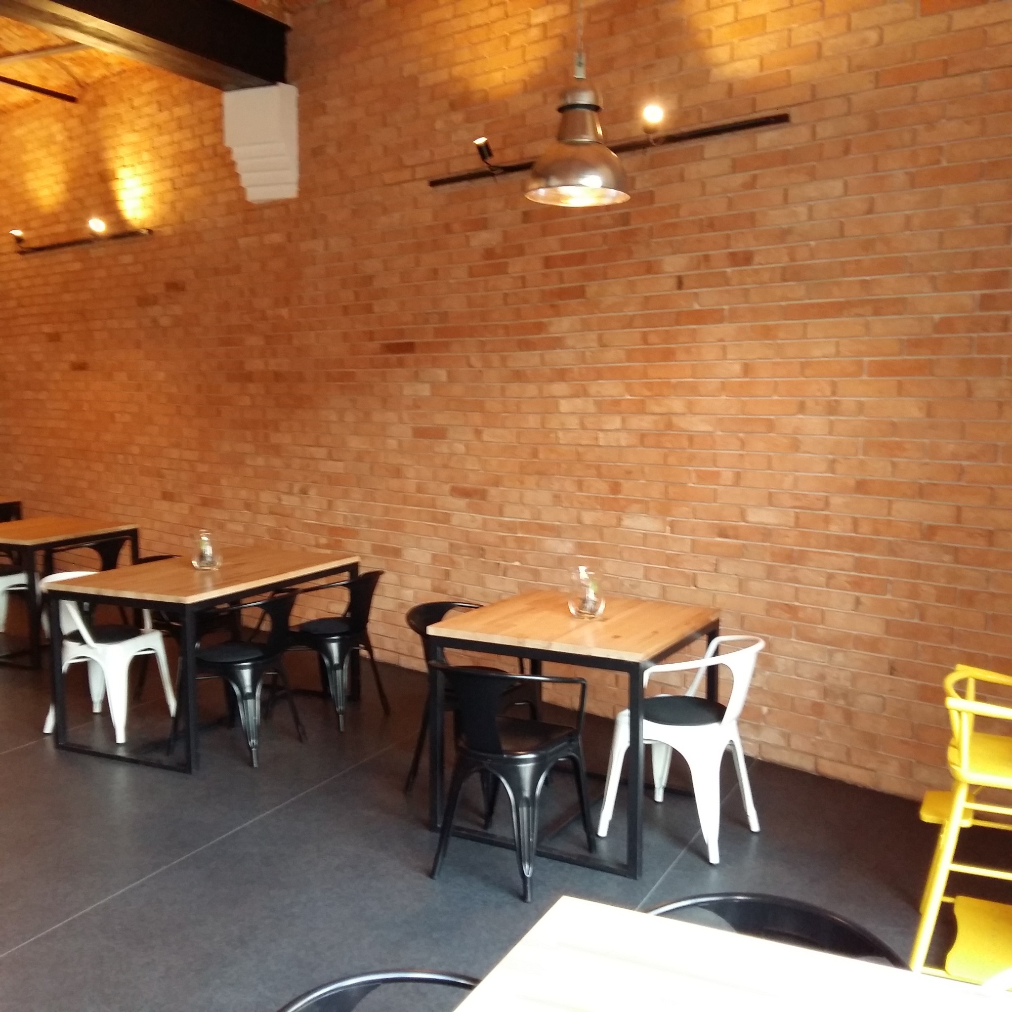 Wnętrze restauracji w stylu loft z ceglaną ścianą, metalowymi lampami wiszącymi i stołami z drewnianymi blatami oraz różnokolorowymi krzesłami, na podłodze ciemne płytki.