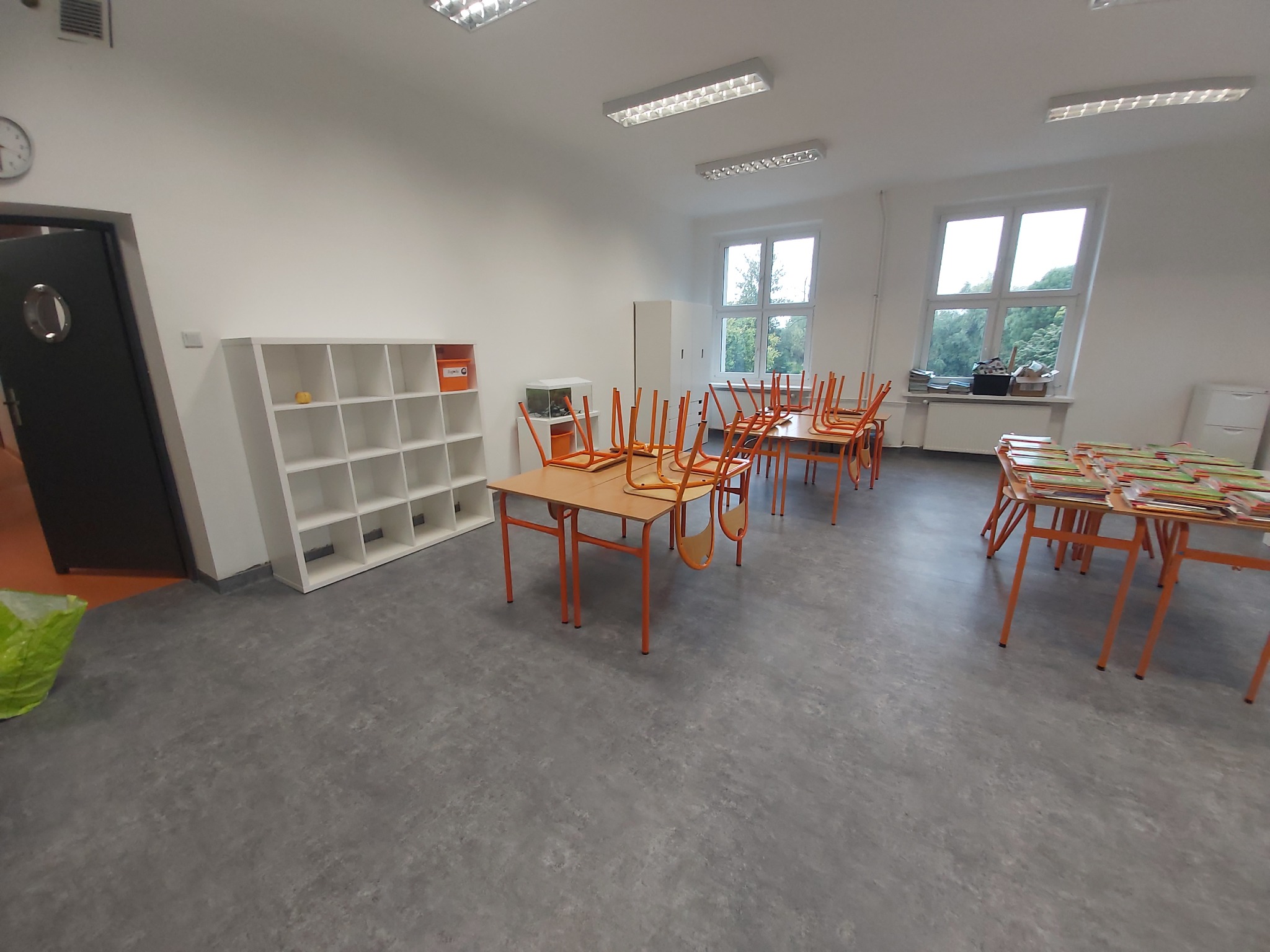 Szara wykładzina w pustej sali lekcyjnej z pomarańczowymi stolikami i krzesłami, białe regały i okna w tle.