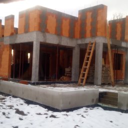 Usługi ogólnobudowlane Patryk Graf - Surowy stan domu murowanego z betonu komórkowego i żelbetu na fundamencie w zimowej scenerii, widoczna drewniana drabina i palety.