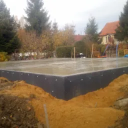 Wylany fundament z hydroizolacją w postaci folii kubełkowej, widoczny teren budowy, dom w tle, pochmurne niebo.