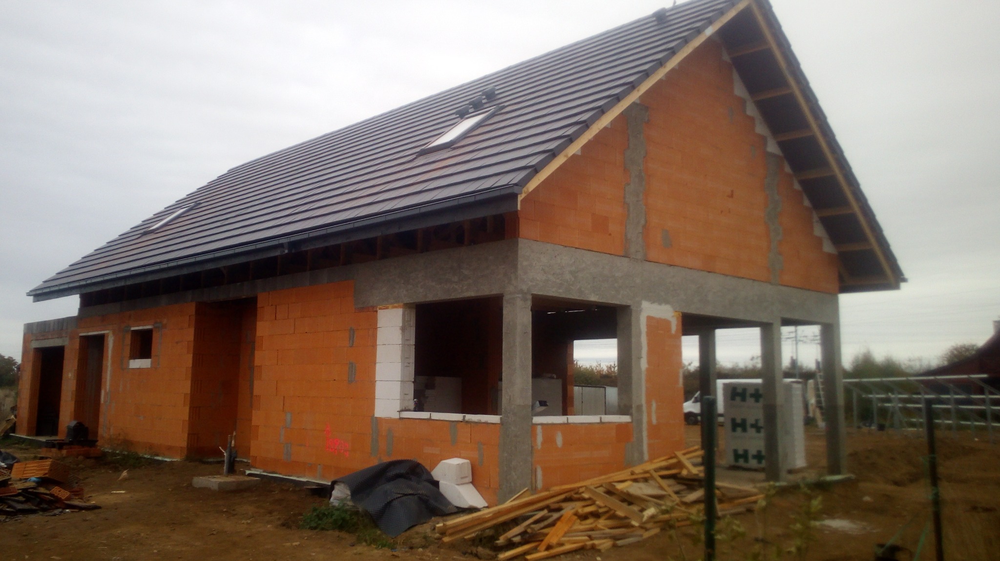 Surowy stan domu jednorodzinnego z czerwonej cegły ceramicznej, widoczne okna z białymi parapetami, dachówka ceramiczna w kolorze antracytowym, szare nadproża betonowe, materiały budowlane na placu...