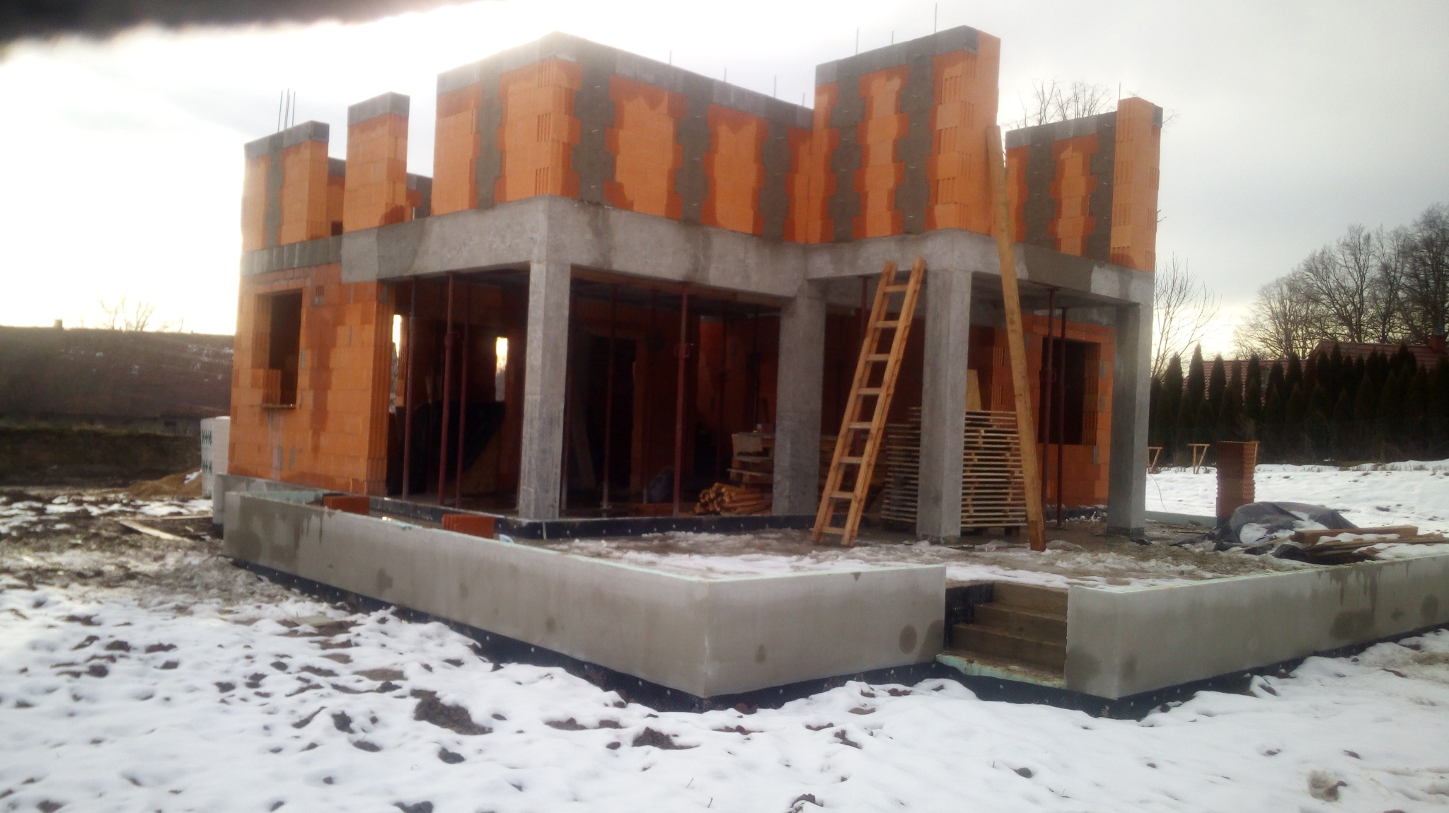 Surowy stan domu murowanego z betonu komórkowego i żelbetu na fundamencie w zimowej scenerii, widoczna drewniana drabina i palety.