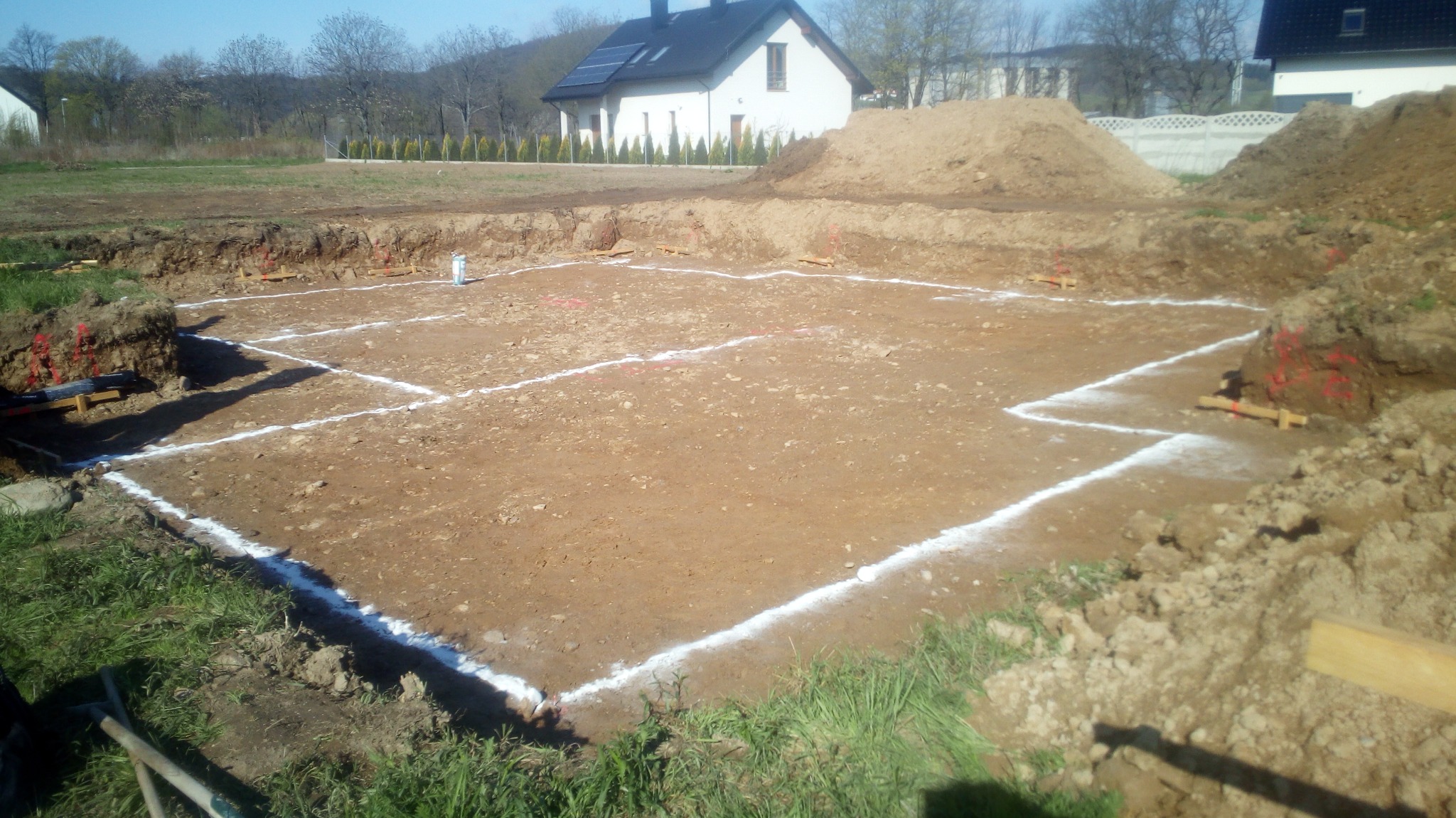 Wytyczony obrys fundamentów domu jednorodzinnego z widocznymi deskami szalunkowymi i stertami ziemi w tle, w oddali biały dom z ciemnym dachem i panelami słonecznymi.