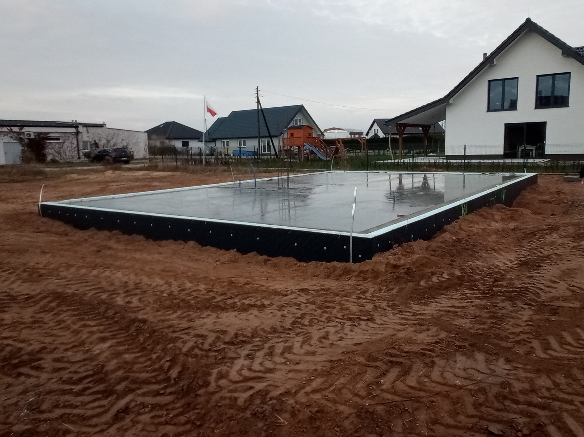 Świeżo wylany fundament płyty betonowej, zabezpieczony czarną folią izolacyjną, z widocznymi prętami zbrojeniowymi i odbiciem nieba na mokrej powierzchni, na tle budynków mieszkalnych i polskiej...