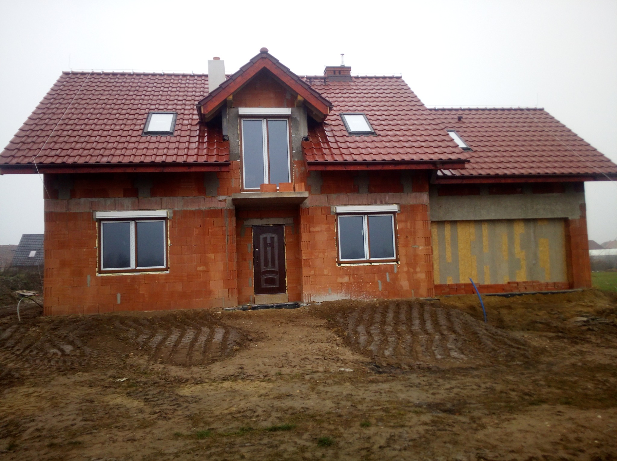 Niedokończony, dwukondygnacyjny budynek z czerwonej cegły z czerwoną dachówką, widoczne okna z białymi ramami, ciemne drzwi wejściowe i przygotowane podłoże.