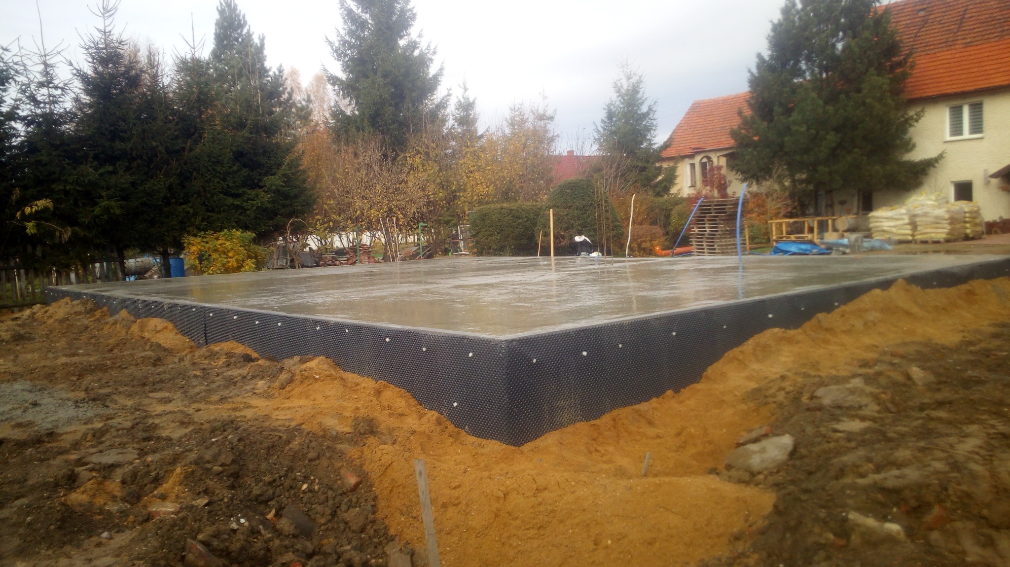 Wylany fundament z hydroizolacją w postaci folii kubełkowej, widoczny teren budowy, dom w tle, pochmurne niebo.