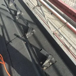 Czarny, bitumiczny dach z zamontowanymi, metalowymi wspornikami pod konstrukcję, widoczne rusztowanie w tle.