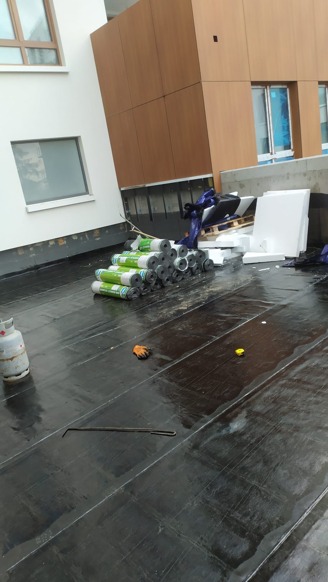 Czarna, błyszcząca powierzchnia dachu pokryta papą termozgrzewalną, z widocznymi rolkami materiału izolacyjnego, butlą gazową, rękawicą roboczą i miarką, na tle fragmentu budynku z oknami...