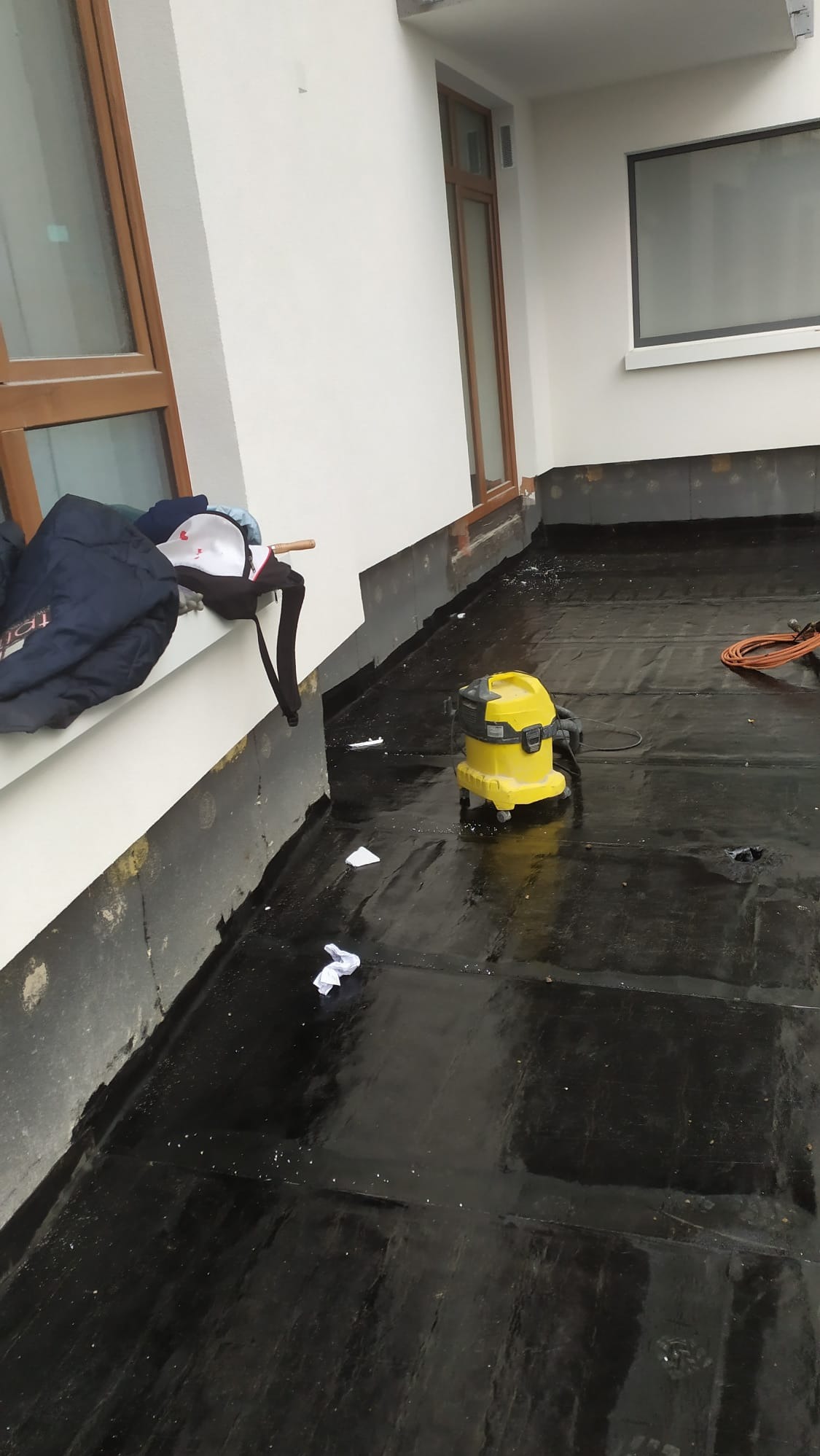 Płaski dach pokryty czarną membraną bitumiczną, widoczny żółty odkurzacz przemysłowy, resztki materiału izolacyjnego, oraz ubrania leżące na parapecie okna.