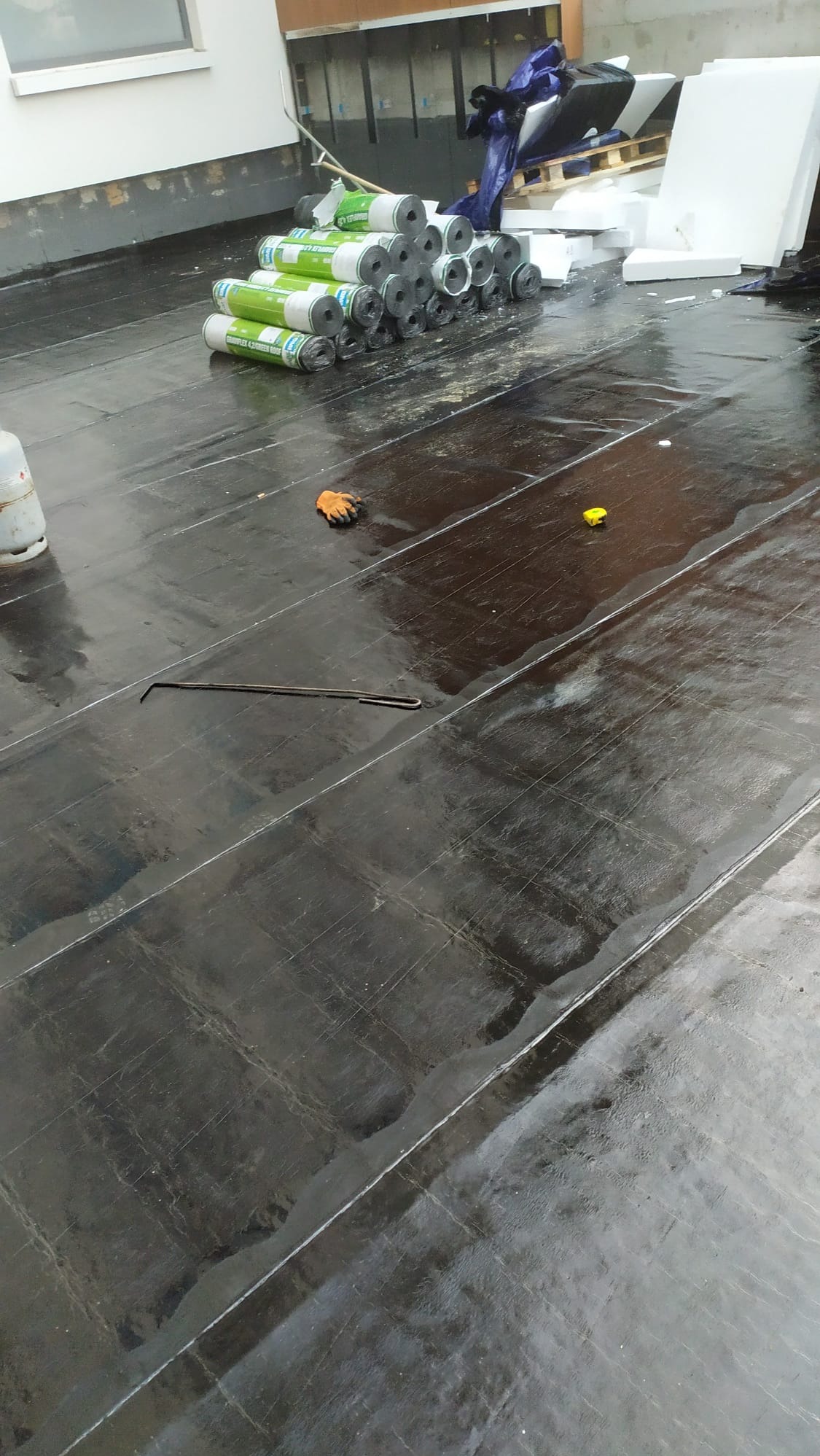 Przygotowanie do izolacji: czarna, błyszcząca powierzchnia pokryta papą, obok zwinięte rolki materiału izolacyjnego, płyty styropianowe, butla gazowa, rękawica robocza i miarka.