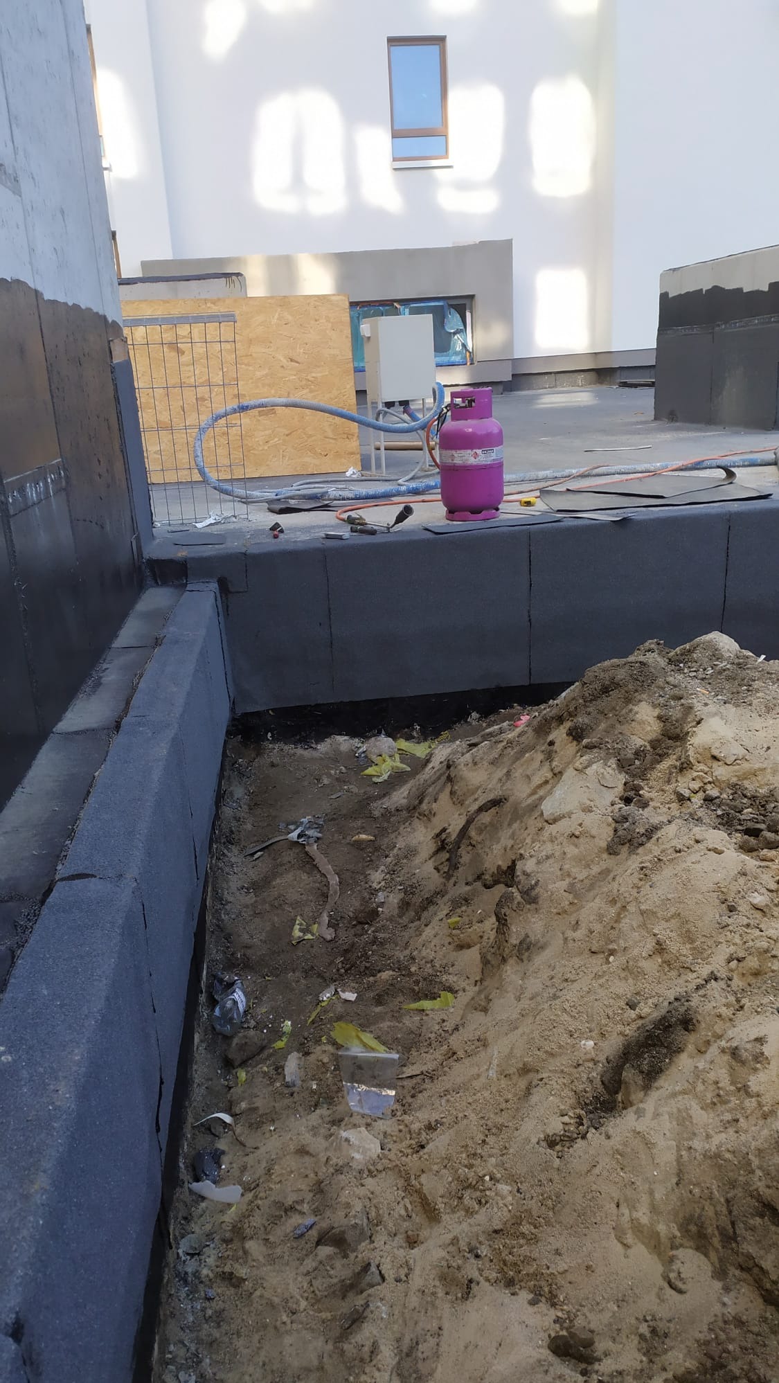 Fundament budynku w trakcie izolacji, widoczna czarna izolacja pionowa, różowa butla gazowa i wykop z ziemią.