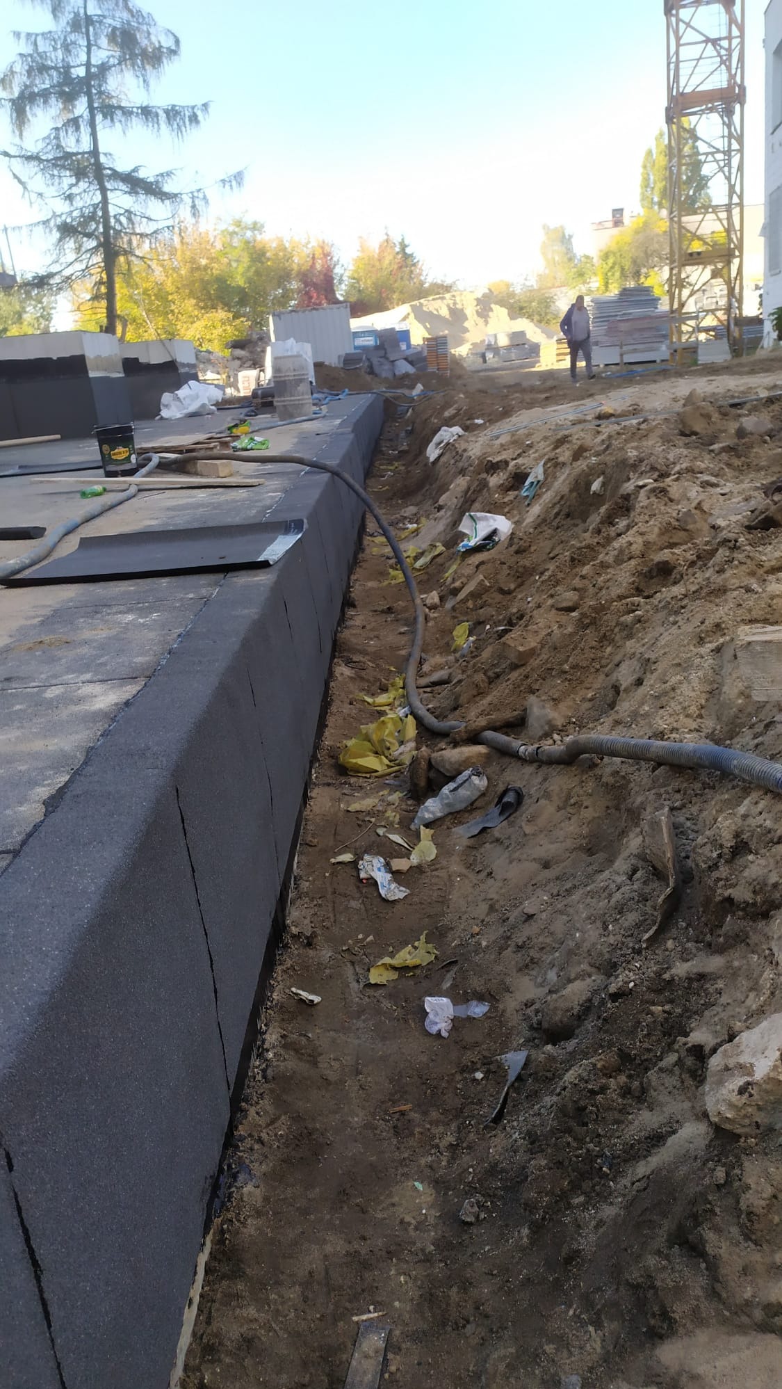 Fundament budynku w trakcie izolacji, widoczne płyty izolacyjne z czarnego styropianu, wykop, gruz, śmieci i węże instalacyjne. W tle budowa i pracownik w oddali.