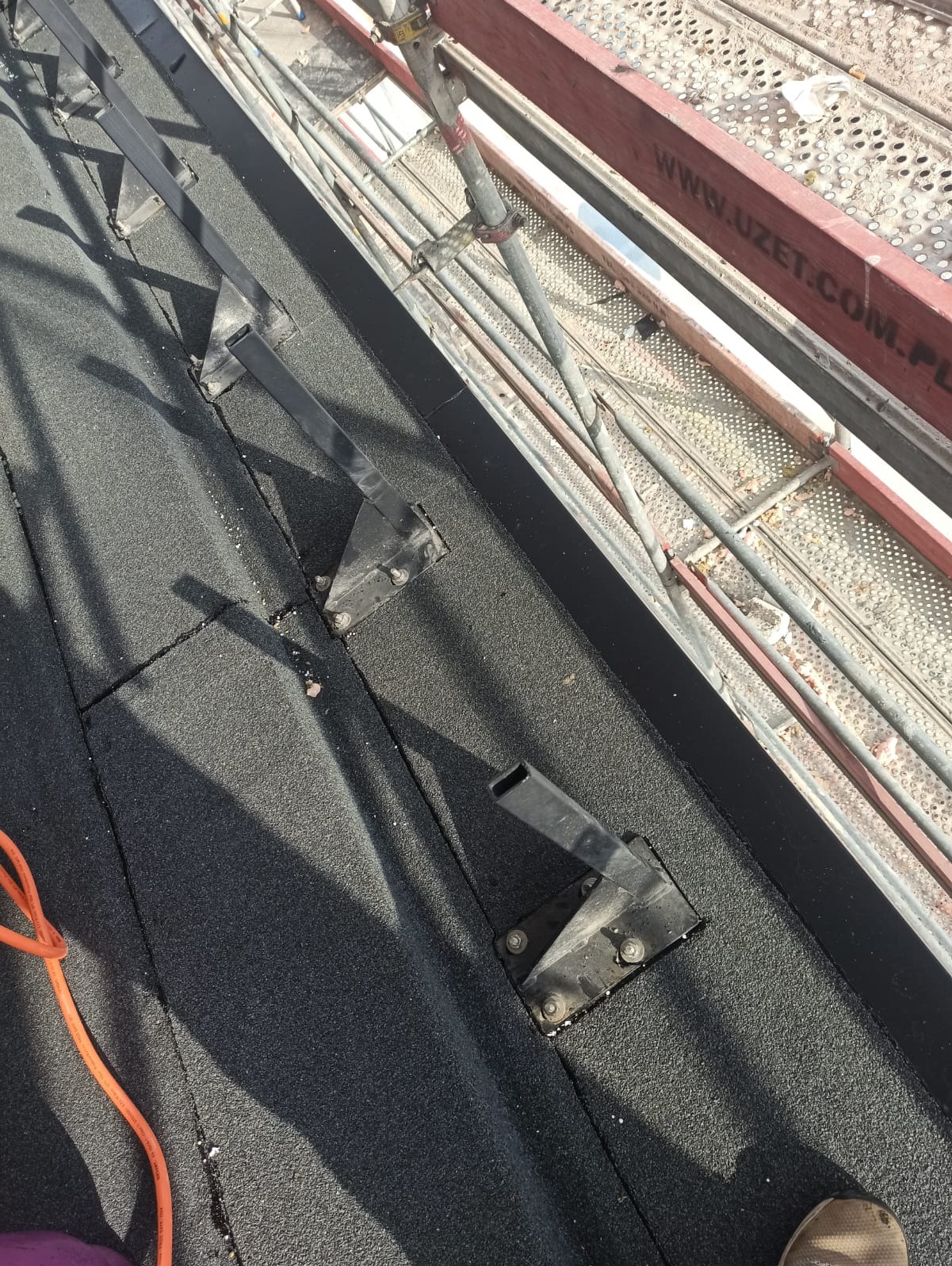 Czarny, bitumiczny dach z zamontowanymi, metalowymi wspornikami pod konstrukcję, widoczne rusztowanie w tle.