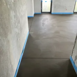 Gładka, szara powierzchnia świeżo wylanej podłogi w surowym wnętrzu z oknami, zabezpieczona niebieską taśmą przy ścianach. Widok z perspektywy wejścia.