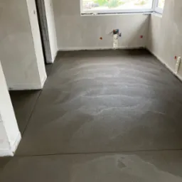 Świeżo wylana, mokra posadzka jastrychowa w jasnym pomieszczeniu z oknem. Widoczne ślady po poziomowaniu i dylatacje. Ściany surowe, przygotowane do dalszych prac.