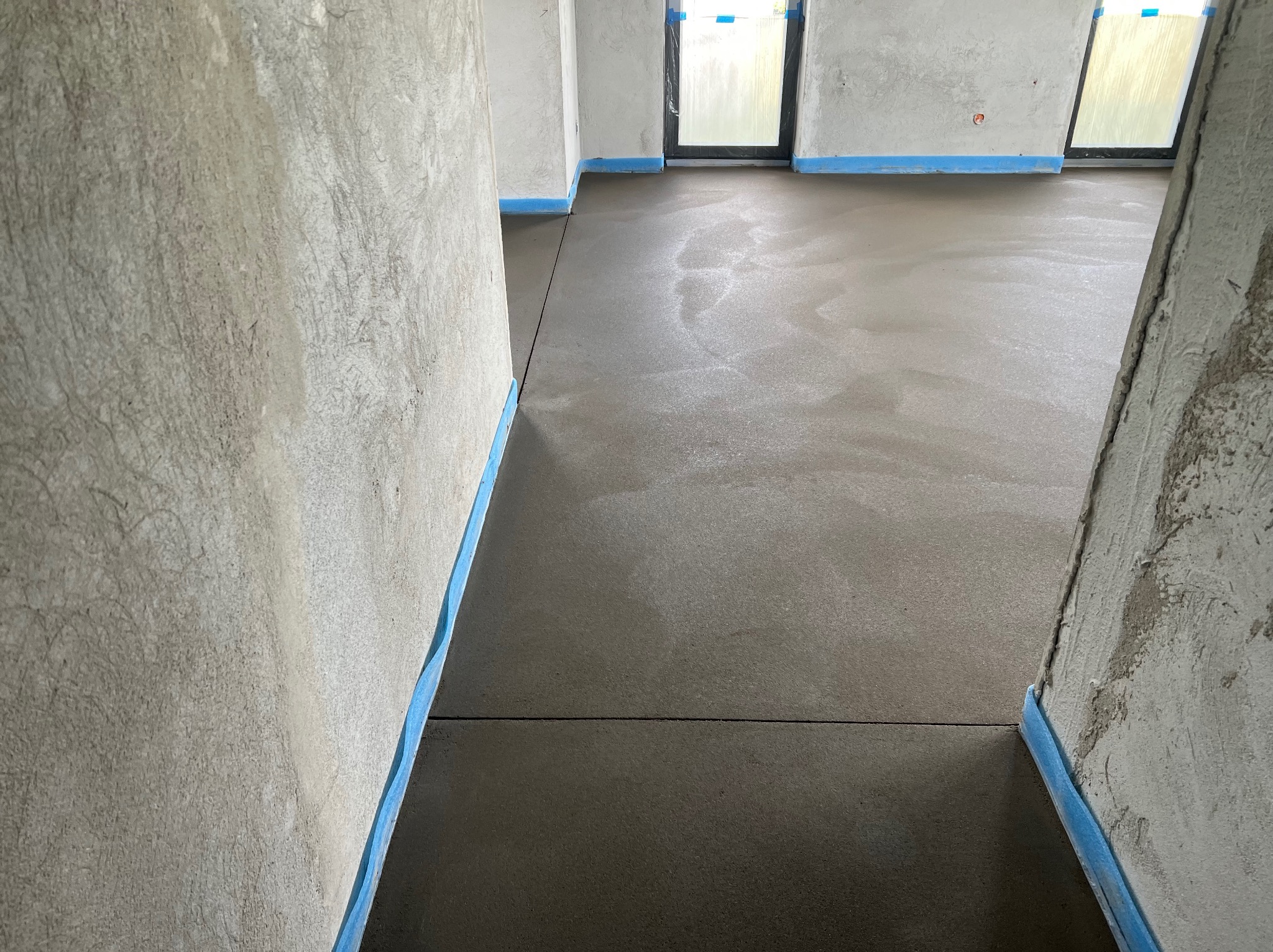 Gładka, szara powierzchnia świeżo wylanej podłogi w surowym wnętrzu z oknami, zabezpieczona niebieską taśmą przy ścianach. Widok z perspektywy wejścia.