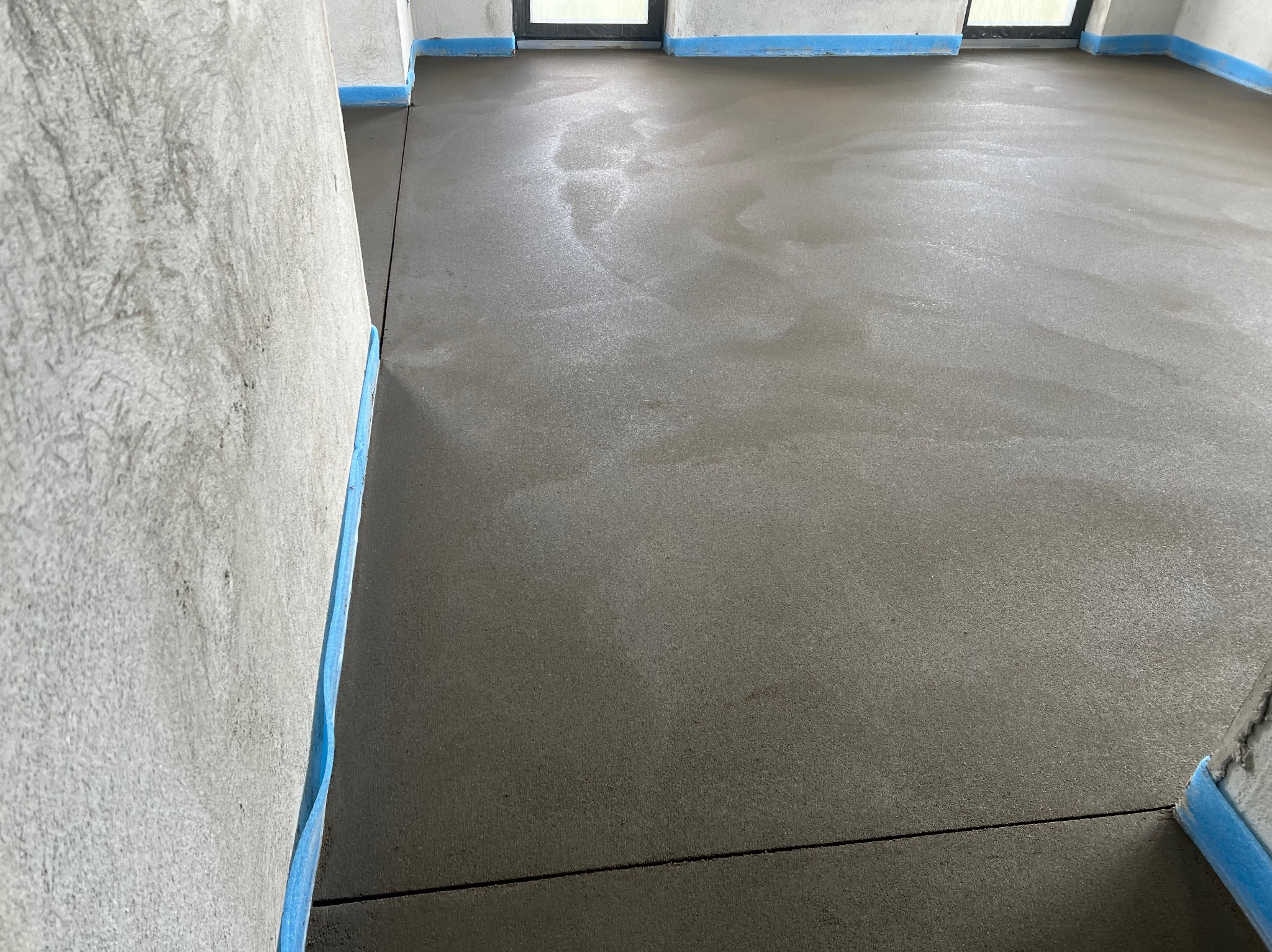 Świeżo wylana, gładka posadzka w pomieszczeniu z zabezpieczonymi ścianami. Widoczne dylatacje i delikatne refleksy światła na powierzchni.