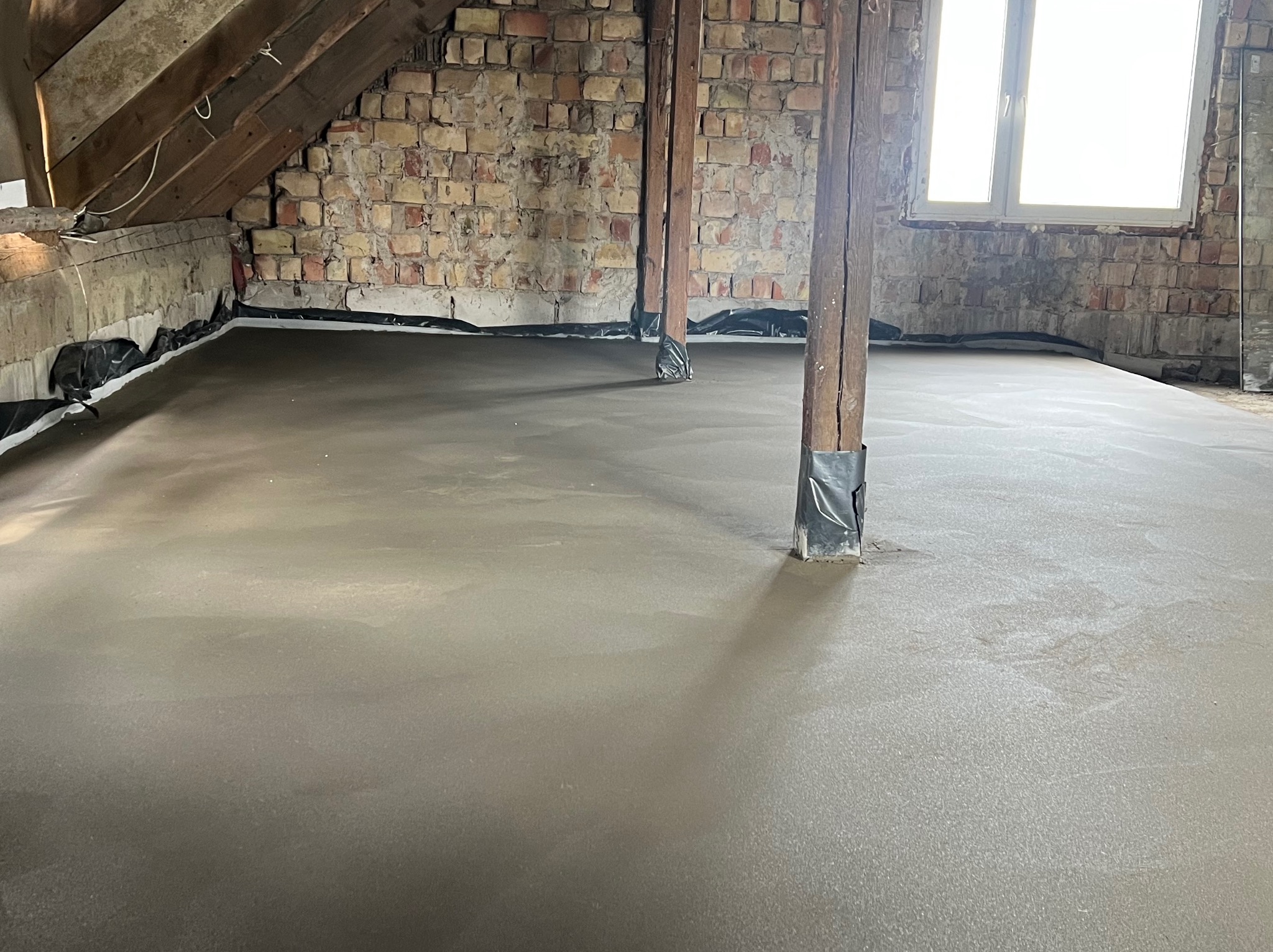 Świeża, gładka wylewka na poddaszu z odsłoniętą cegłą, drewnianymi belkami stropowymi i słupami. Folia wzdłuż ścian. Okno w tle.