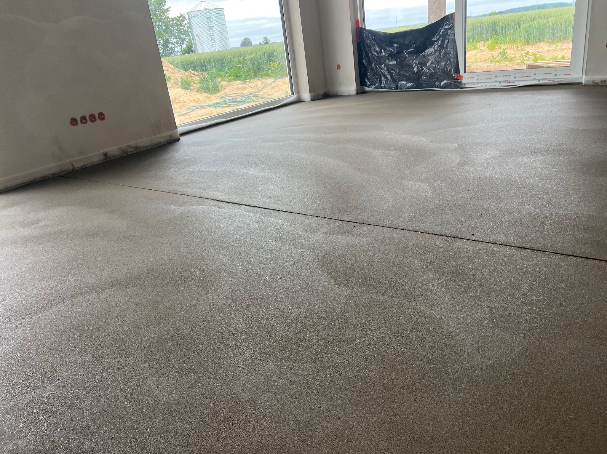 Świeżo wylana, gładka posadzka w jasnym pomieszczeniu z dużymi oknami, przez które widać pole i silos. Przewody elektryczne wystają ze ściany.