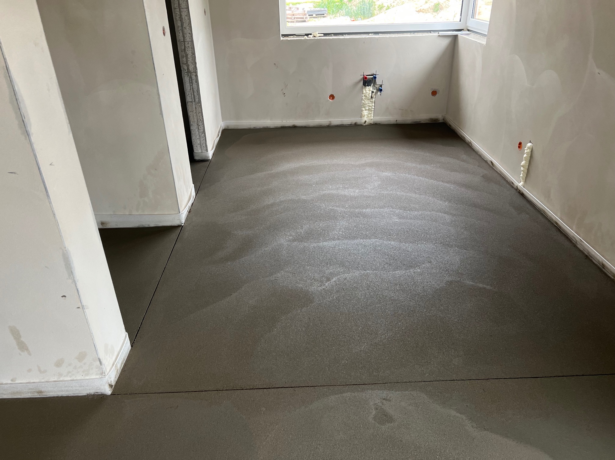 Świeżo wylana, mokra posadzka jastrychowa w jasnym pomieszczeniu z oknem. Widoczne ślady po poziomowaniu i dylatacje. Ściany surowe, przygotowane do dalszych prac.
