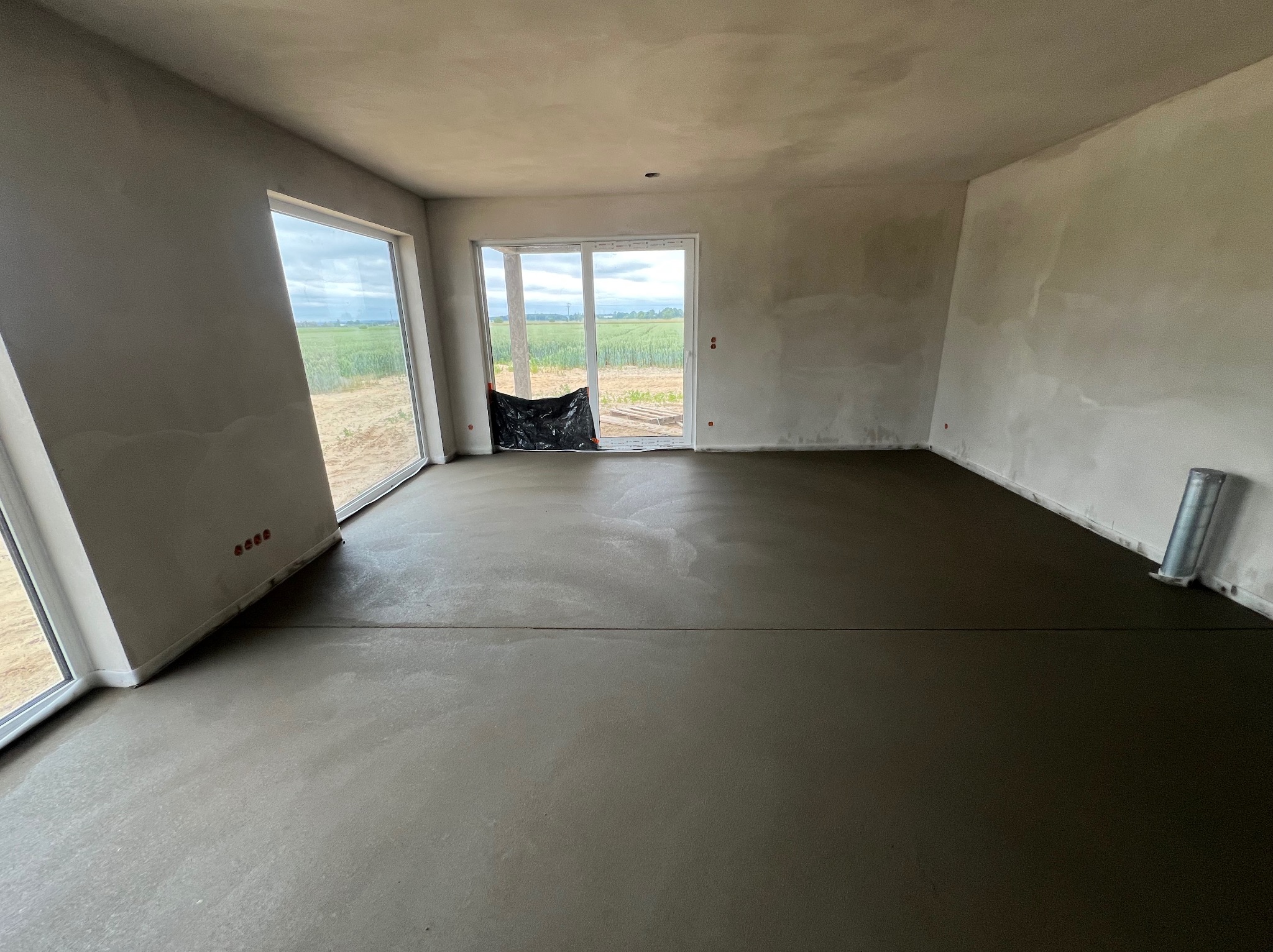 Surowe wnętrze z wylaną, gładką posadzką, widok na zielone pole przez okna. Ściany w stanie surowym, przygotowane do dalszych prac wykończeniowych.
