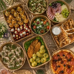 RUKOLA BARBARA MICHALIK - Różnorodne dania w ekologicznych pojemnikach: pizza z pepperoni i oliwkami, sałatki z orzechami i fetą, szaszłyki caprese, pieczone ziemniaki z kurczakiem, wędliny i sery. Aranżacja na drewnianym...