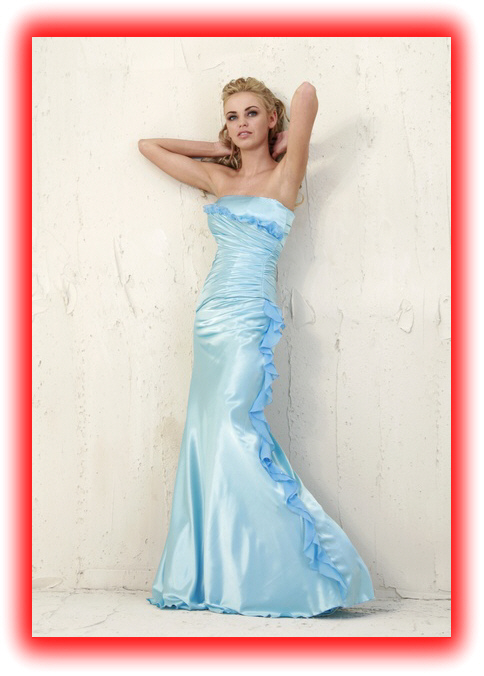 Modelka w błękitnej, satynowej sukni balowej bez ramiączek, z falbanami, oparta o białą ścianę z fakturą.