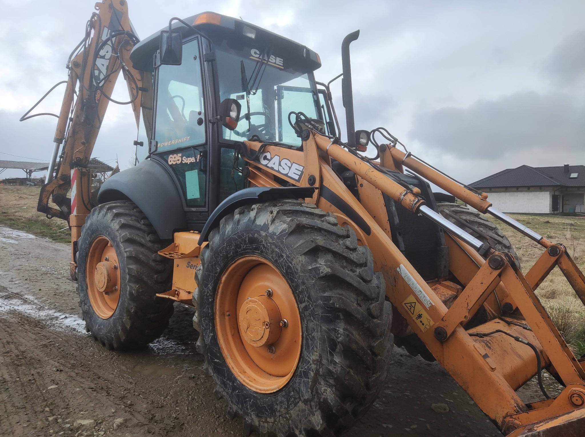 Pomarańczowa koparko-ładowarka CASE 695 Super I na błotnistym terenie z widocznym fragmentem budowanego domu w tle.