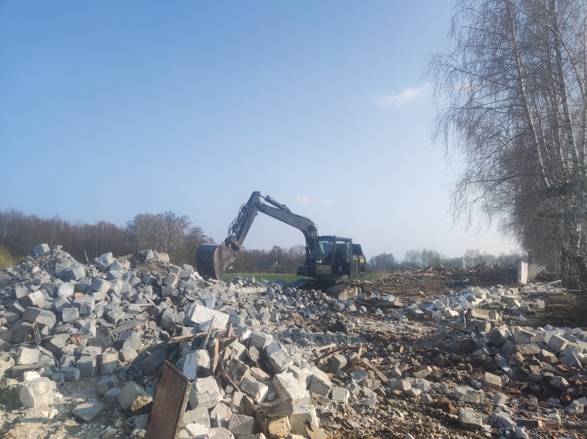 Ciężki sprzęt - koparka stoi na gruzowisku po rozbiórce budynku, z fragmentami muru porozrzucanymi wokół, w tle drzewa bez liści i błękitne niebo.