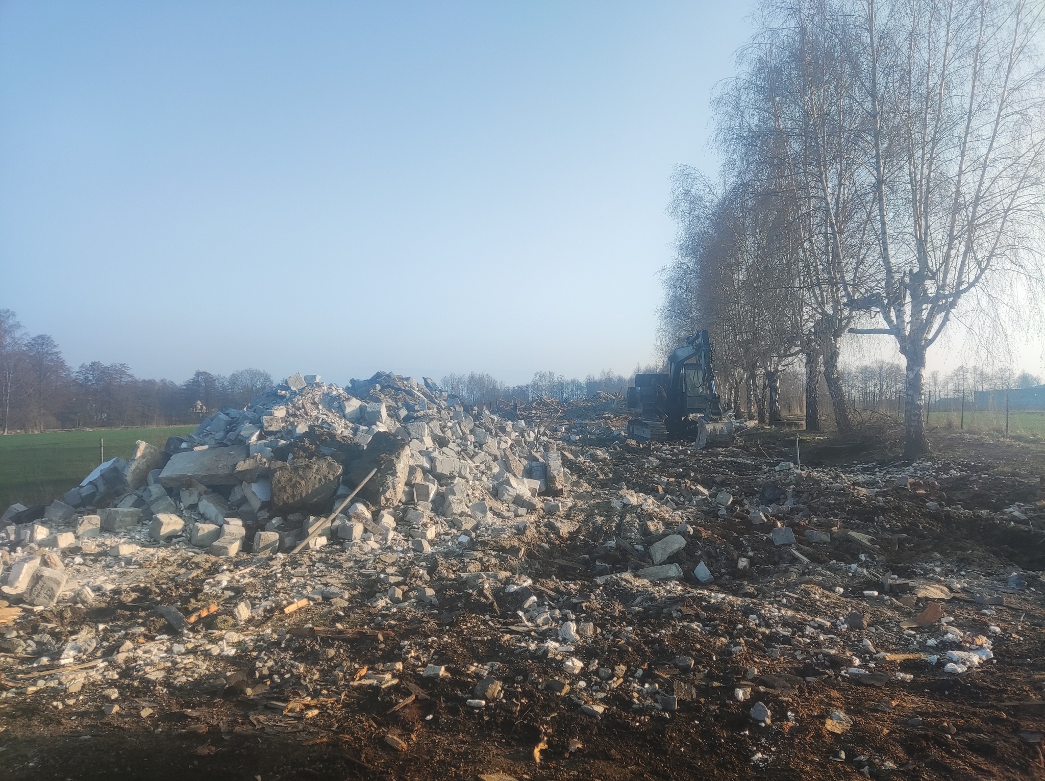 Kupa gruzu po rozbiórce budynku, z widoczną koparką w tle i rzędem drzew bez liści po prawej stronie, sfotografowana w jasny, słoneczny dzień.