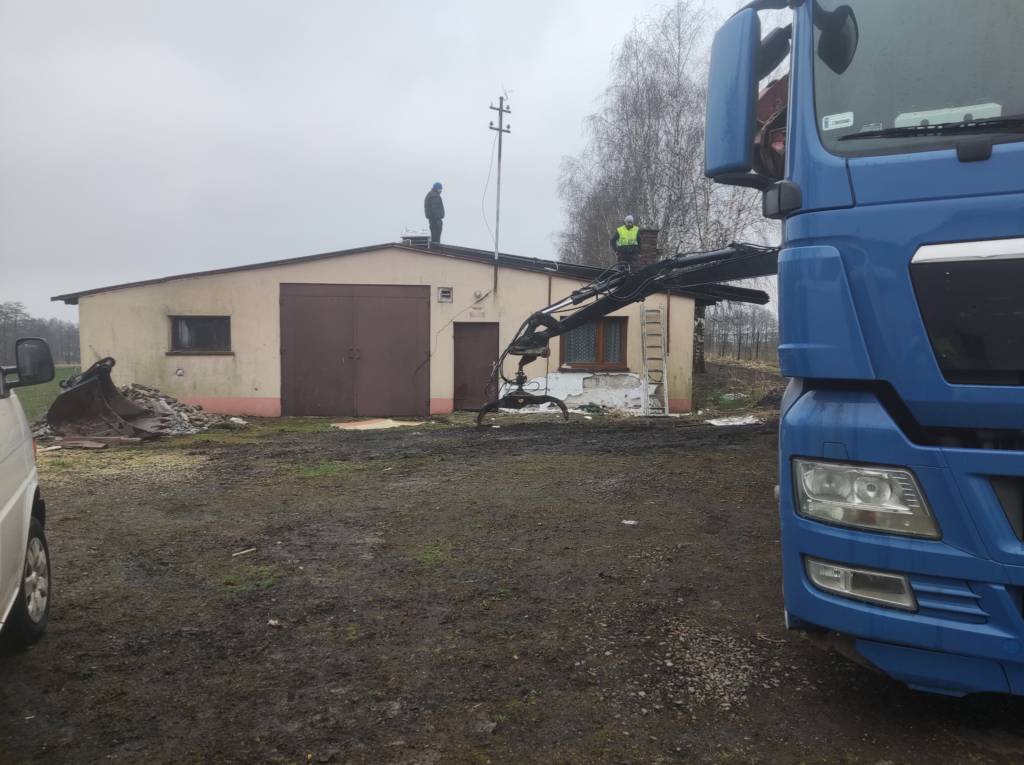 Dwóch robotników na dachu jednopiętrowego budynku z cegły, przygotowujących się do demontażu, widoczny dźwig samochodowy i gruzowisko w pobliżu.