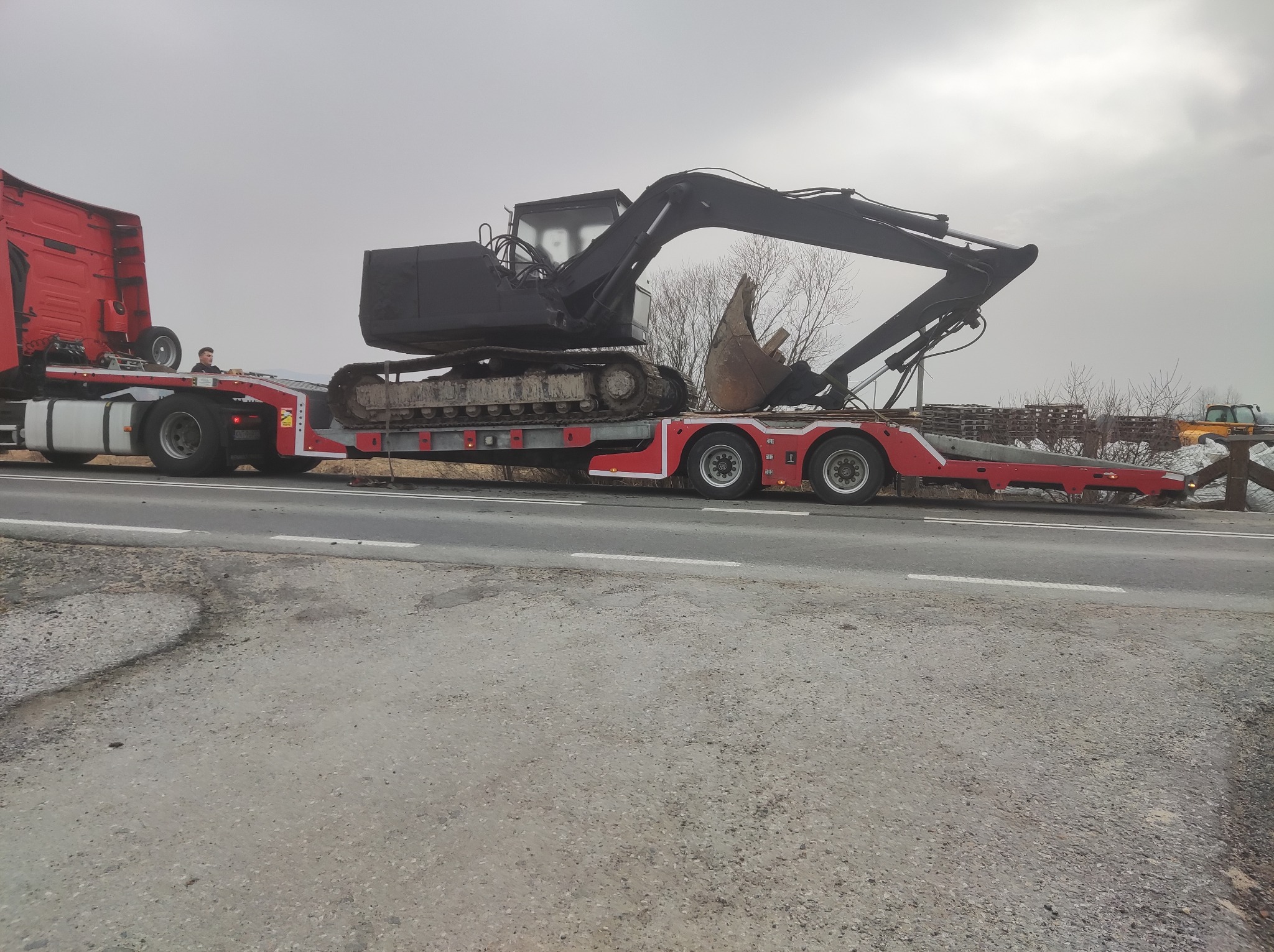 Czarny, brudny od ziemi, gąsienicowy pojazd budowlany, prawdopodobnie koparka, transportowany na niskopodwoziowej platformie ciężarówki, widok z boku na tle szarego nieba i pobocza drogi.