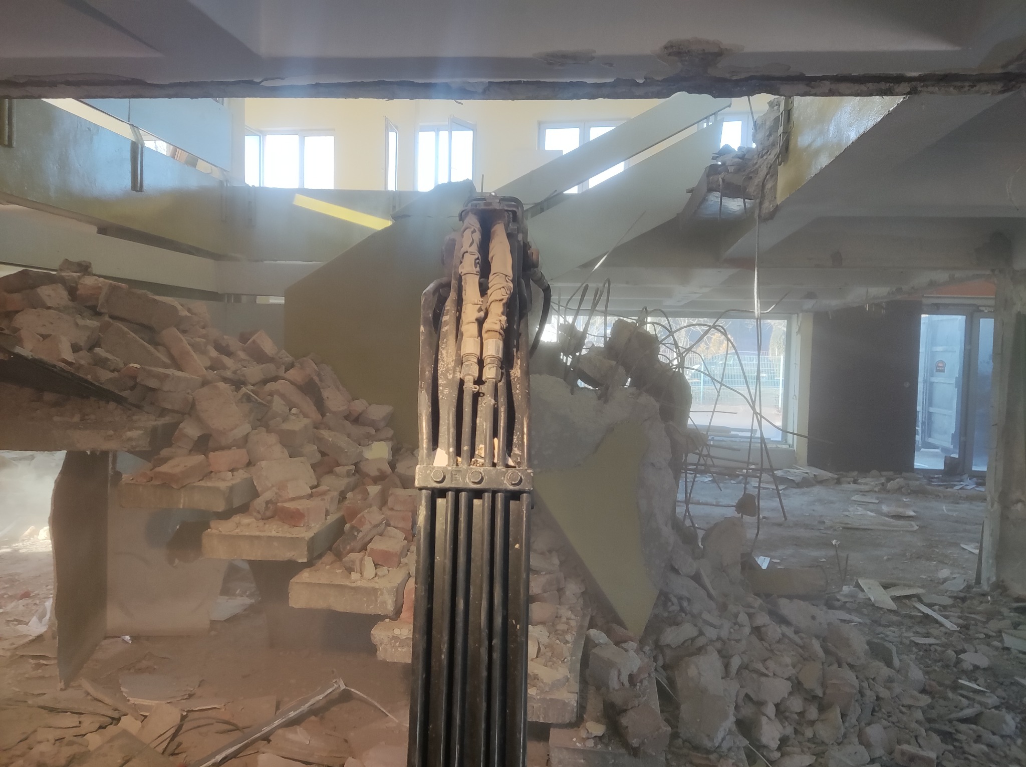 Hydrauliczne nożyce do betonu zamontowane na koparce w trakcie wyburzania schodów wewnątrz budynku, gruz ceglany i betonowy na podłodze, widoczne okna w tle.