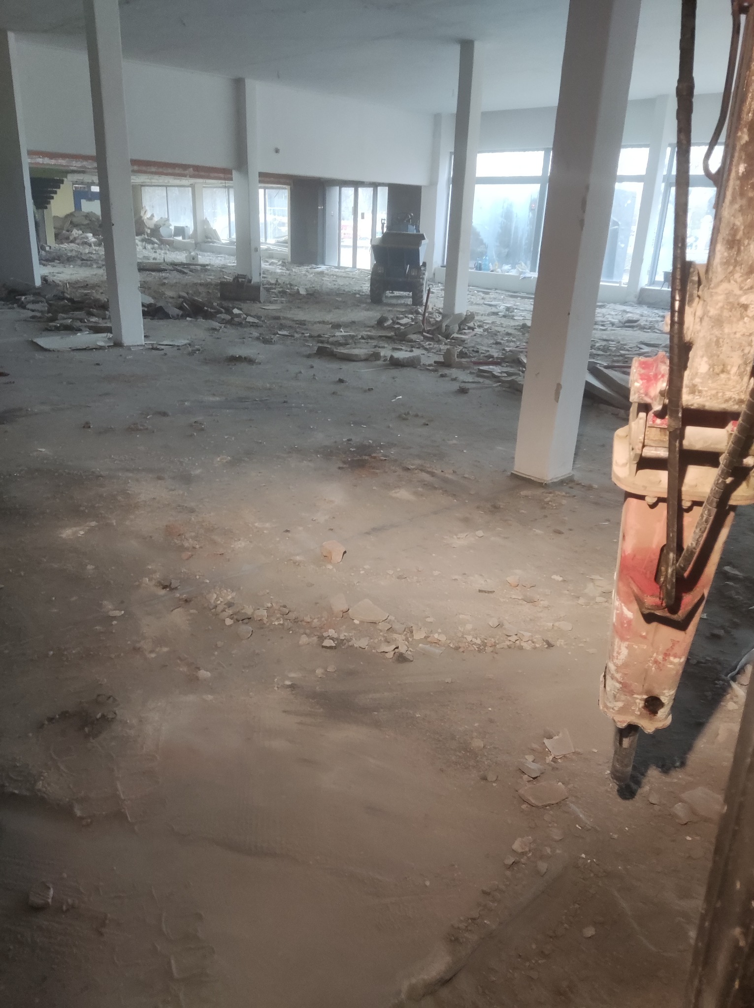 Wnętrze budynku po rozbiórce, widoczne gruz, odsłonięte elementy konstrukcyjne, w tle maszyna budowlana oraz fragment hydraulicznego młota wyburzeniowego w pierwszym planie, oświetlenie naturalne...